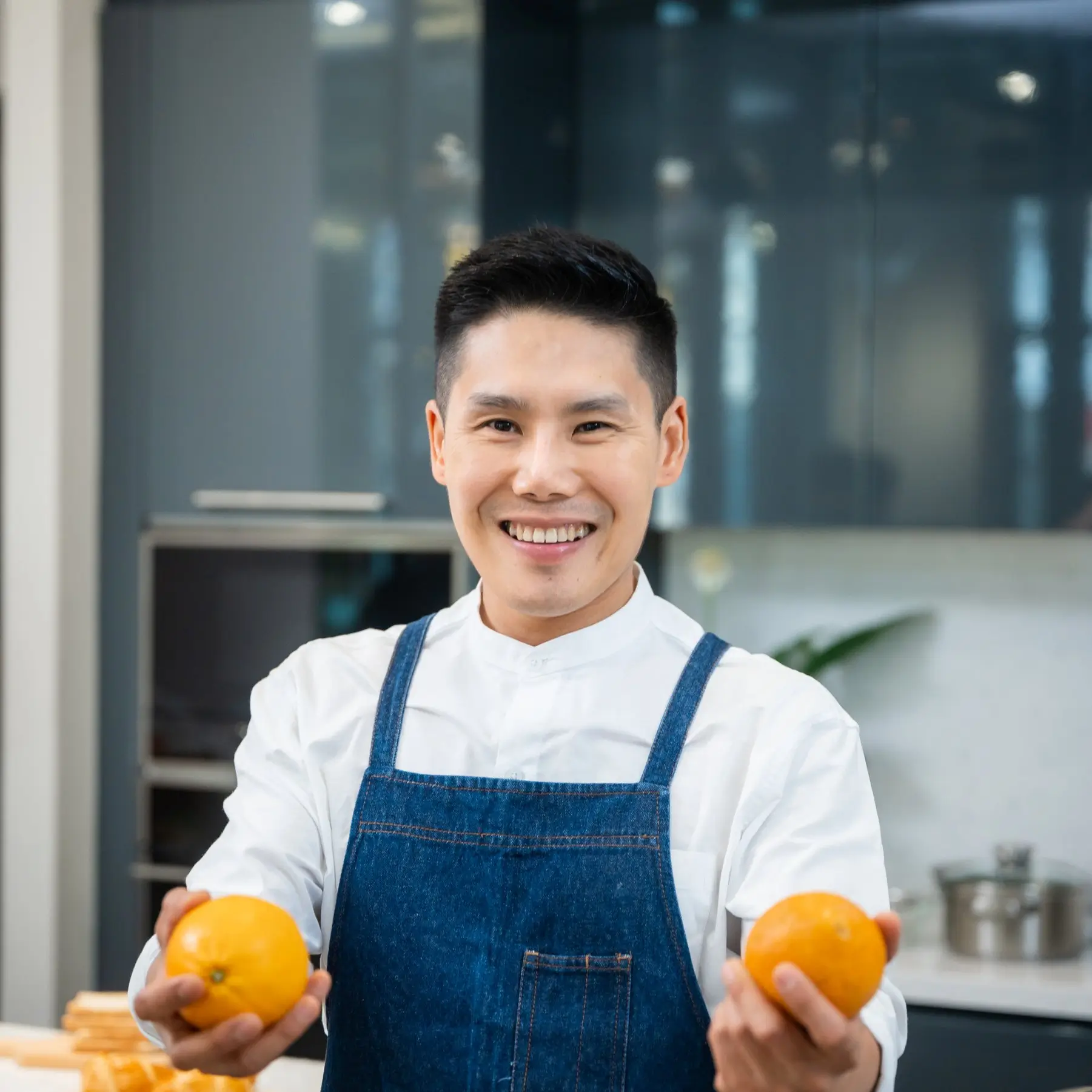 Portrait of Ethan Park, Kitchen Assistant and Recipe Tester at SliceSync, smiling in a modern kitchen while holding two oranges.