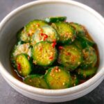 A bowl of Korean cucumber salad with sliced cucumbers in a glossy spicy dressing, sprinkled with sesame seeds and red chili pieces.