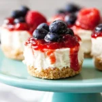 No-Bake Mini Cheesecakes topped with blueberries, raspberries, and berry sauce on a cake stand